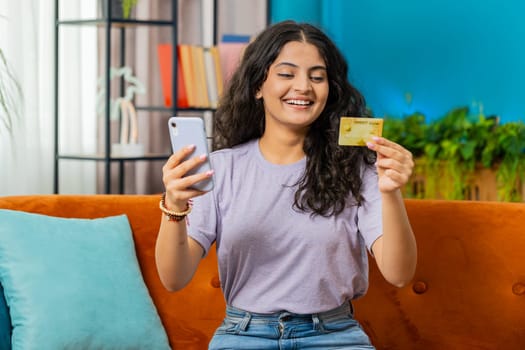 Indian woman use credit bank card and smartphone while transferring money, purchases online shopping cashless order food delivery at home apartment indoors. Happy Arabian girl sits in room on couch