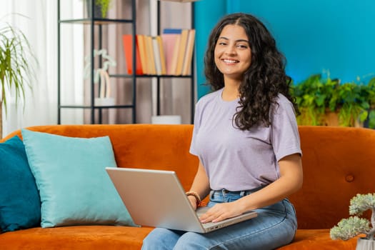 Happy Indian woman sitting on couch after finishing laptop work in living room. Smiling Arabian girl freelancer works online remote job at home sofa. E-learning, browsing internet on notebook computer