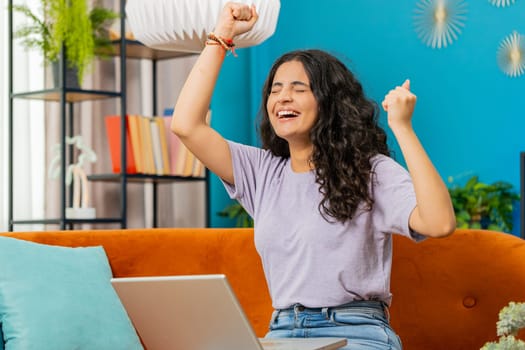 Surprised Indian young woman using laptop computer, receive mail good news message shocked by sudden victory celebrate lottery jackpot win purchases online shopping play game at home apartment on sofa