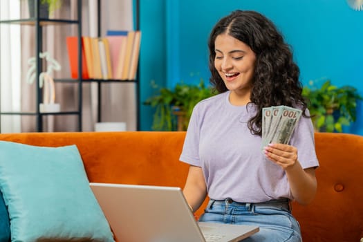 Planning budget. Rich happy Indian woman counting money cash use laptop computer calculate domestic bills at home. Arabian girl satisfied of income earnings, saves money for planned vacation, gifts