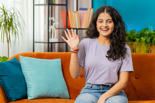 Hello. Indian happy woman smiling friendly at camera and waving hands gesturing hello hi, greeting or goodbye welcoming with hospitable expression at home. Arabian girl sitting on sofa in living room