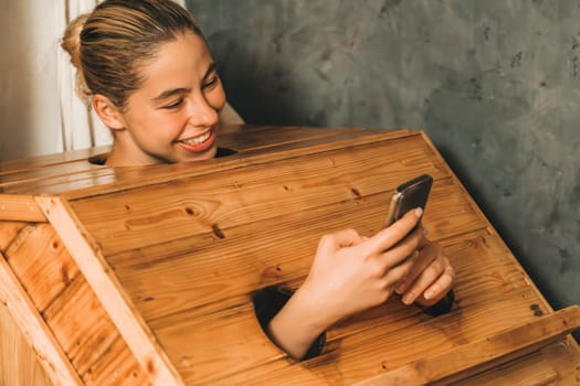 A portrait of gorgeous caucasian woman playing her mobile phone while using wooden sauna cabinet in warm tone. Attractive female with beautiful skin taking a photo. Gray background. Tranquility