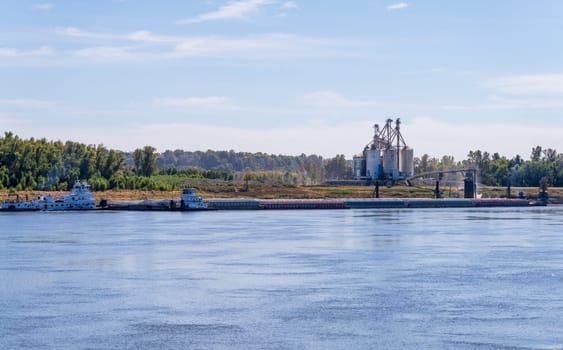 Tugboat pushing freight barges past grain loading dock in farming country in Kentucky