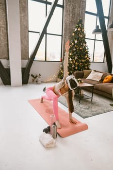 The stylish young woman, in her workout outfit and a virtual reality headset, does yoga poses beside a Christmas tree. High quality photo