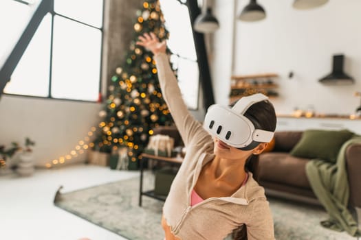 A fitness-oriented young woman practices yoga poses while wearing VR glasses by a Christmas tree. High quality photo