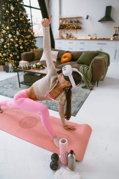 Wearing workout clothes and a virtual reality headset, the fashionable young woman engages in yoga exercises next to a Christmas tree. High quality photo