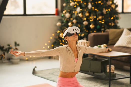 In athletic clothing, the chic young woman exercises yoga using a virtual reality headset beside a Christmas tree. High quality photo