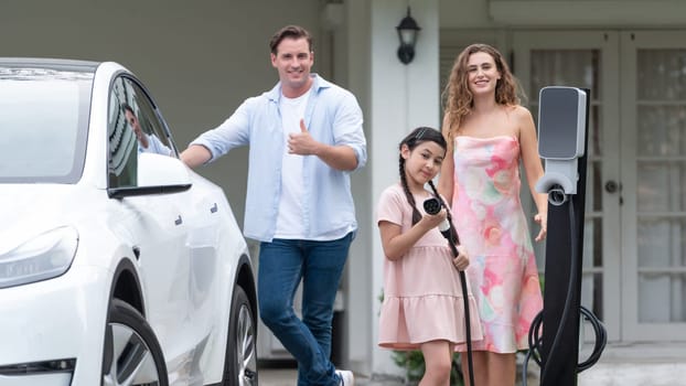 Modern family with environmental conscious recharging EV car with home EV charging station, Happy family with little young girl holding and pointing EV charger at camera. Synchronos