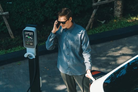 Young man travel with EV electric car charging in green sustainable city outdoor garden in summer shows urban sustainability lifestyle by green clean rechargeable energy of electric vehicle innards