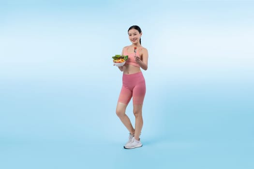 Young sporty Asian woman in sportswear holding salad bowl fill with vibrant of fruit and vegetable. Natural youthful and fit body lifestyle with balance nutrition on isolated background. Vigorous