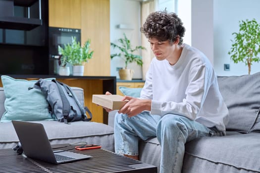 Young male unpacking an online purchase, sitting on couch at home. Internet online shopping, delivery, youth concept