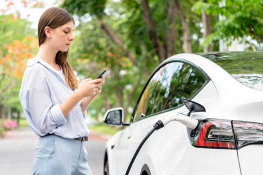 Young woman recharge EV electric vehicle battery from EV charging station and using smartphone online banking to pay for electricity in city park. Eco friendly vehicle travel with EV car. Exalt