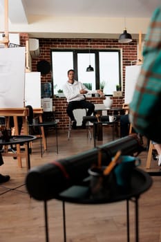 Young African American man teacher sitting in classroom in from of students, talking about fundamentals of still life drawing during group art class. Prooffesion male artist teaching sketching lesson