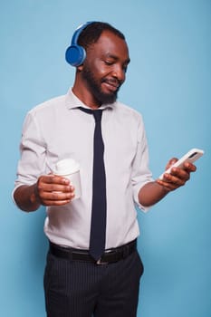 Busy black man with wireless headset browsing social media on mobile device while holding a beverage. Office worker on break listening to online music while drinking his cup of coffee.