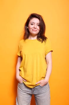 In studio, excited female person stands in front of isolated orange background. Joyful woman poses confidently, casually looking at the camera. Stylish adult with hands in pockets and a smile.