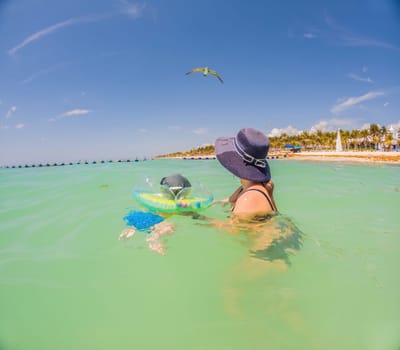 Adorable baby swims in an inflatable ring around his neck, enjoying the sea with his mom