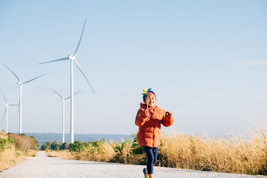 Little girl's happiness near windmills holding pinwheels embodies playful wind energy education. Clean electricity's beauty showcased in a cheerful sunny wind turbine setting.
