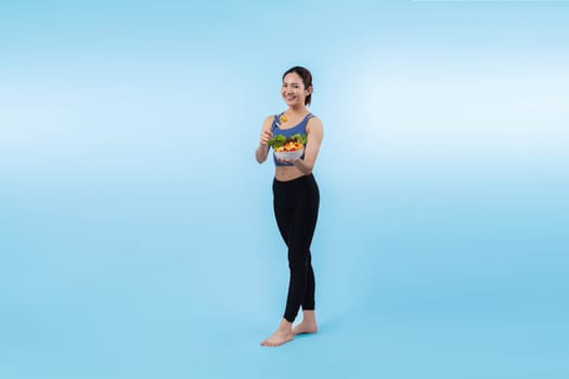 Young sporty Asian woman in sportswear holding salad bowl fill with vibrant of fruit and vegetable. Natural youthful and fit body lifestyle with balance nutrition on isolated background. Vigorous