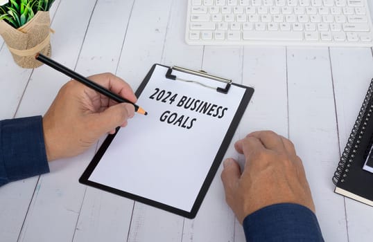 Hands writing a 2024 business goals on a clip board on white wooden desk. Business and finance concept.