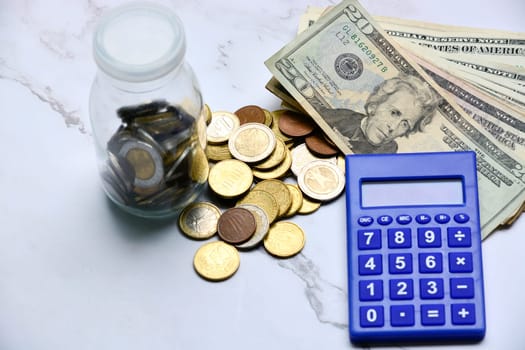 Glass jar with multicurrency coins, US dollar bills and calculator white table. Saving and travelling concept