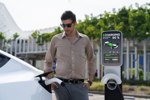 Young man put EV charger to recharge electric car's battery from charging station in city commercial parking lot. Rechargeable EV car for sustainable environmental friendly urban travel. Expedient