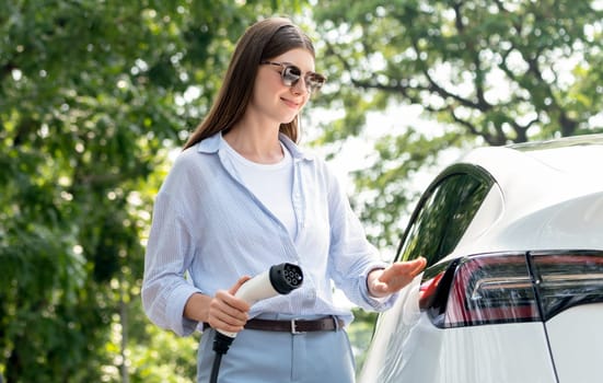 Young woman recharging battery for electric car during road trip travel EV car in natural forest or national park. Eco friendly travel during vacation and holiday. Exalt