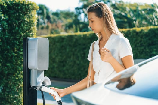 Young woman travel with EV electric car charging in green sustainable city outdoor garden in summer. Urban sustainability lifestyle by green clean rechargeable energy of electric BEV vehicle innards