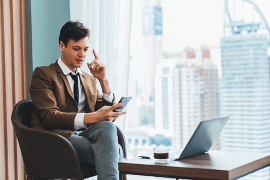 Closeup of handsome businessman using mobile phone while sitting near window with skyscraper view. Professional male leader taking a photo marketing, business project displayed on laptop. Ornamented.