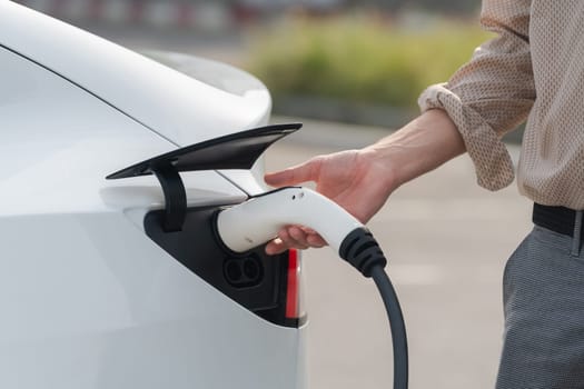 Young man put EV charger to recharge electric car's battery from charging station in city commercial parking lot. Rechargeable EV car for sustainable environmental friendly urban travel. Expedient