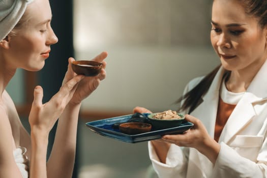 A beautiful young woman in white towel smells herbal facial mask. Professional therapist holding tray contained homemade mask at spa salon, bathroom surrounded by aroma essence. Close up. Tranquility.