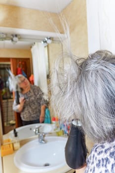 mature white-haired woman drying her hair with a hand-held hair dryer in front of a mirror