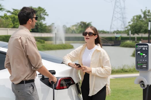 Young couple recharge electric car's battery from charging station in outdoor green city park in springtime. Rechargeable EV car for sustainable environmental friendly urban travel lifestyle.Expedient