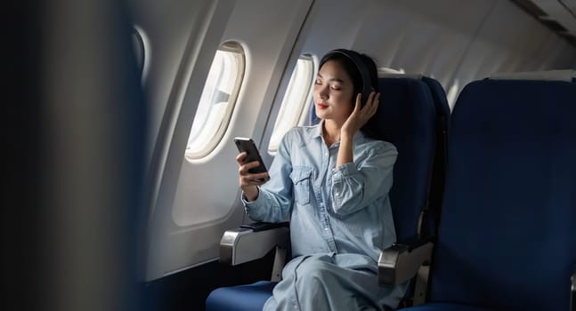 Young female listening song during flight in first class cabin using smartphone, woman entertain on airplane board enjoy music in headphone from smartphone.
