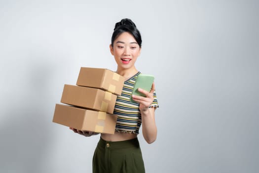 Young happy asian woman wear summer casual clothes hold stack cardboard blank boxes use mobile cell phone 