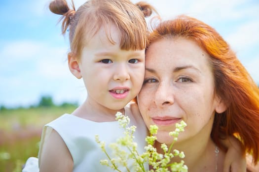 Happy female family with mother and daughter on green and yellow meadow full of grass and flower. Woman with red hair and blonde girl having fun, joy and hug in sunny summer day. Concept family love