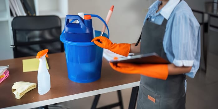 A cleaning woman is standing inside a building holding with a blue tank on the side chemicals and facilities for tidying.