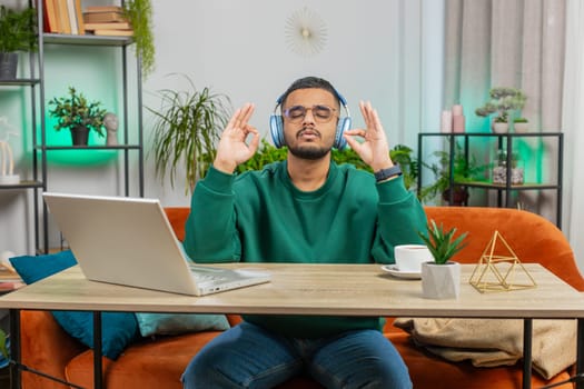 Meditation, relaxing, taking a break. Indian man listening music breathes deeply, eyes closed meditating with concentrated thoughts, peaceful mind. Arabian guy sits at home office room table workplace