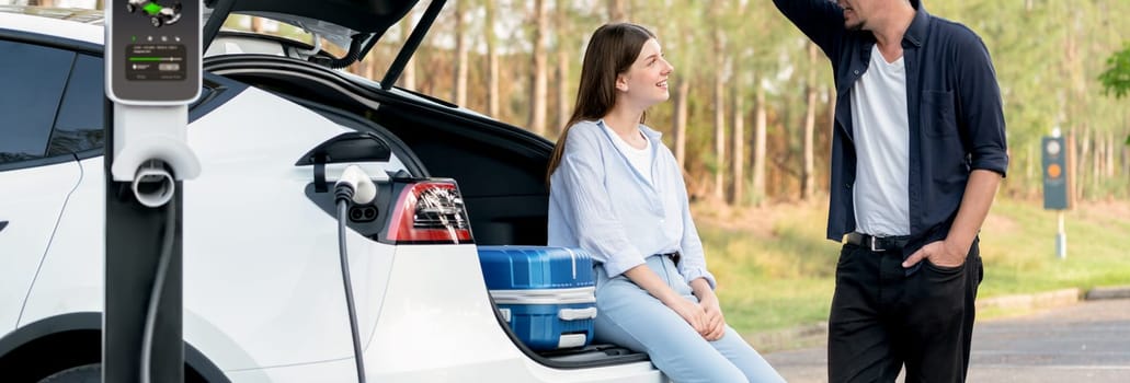 Panorama banner couple recharging battery for electric car while relaxing and sit on the trunk during road trip travel EV car in autumnal forest. Eco friendly travel on vacation during autumn. Exalt