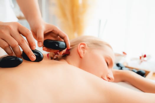 Hot stone massage at spa salon in luxury resort with day light serenity ambient, blissful woman customer enjoying spa basalt stone massage glide over body with soothing warmth. Quiescent