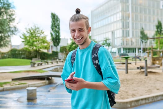 Fashionable cheerful laughing young male with backpack smartphone looking at camera outdoors in city. Hipster with trendy hairstyle beard, mobile applications for tourism travel communication study