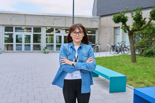 Middle aged confident woman school teacher, mentor, psychologist, counselor, social worker with crossed arms looking at camera near school building, outdoor