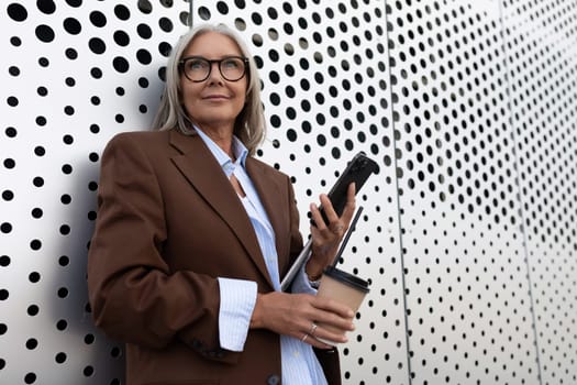 successful gray-haired business grandmother dressed stylishly uses a smartphone while walking along the city street.