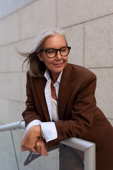 successful free gray-haired business grandmother is dressed in a stylish brown suit against the backdrop of the stairs of the business center.
