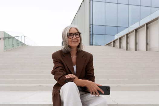 happy mature old lady with gray hair and glasses dressed in a stylish jacket walks around the city and sits on the stairs in the city.