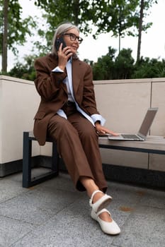 mature business woman with gray hair dressed in a brown suit is holding an online meeting using a laptop in the street.