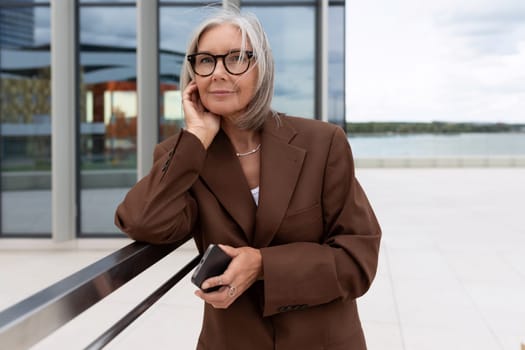 a slender senior business woman with gray hair dressed in an elegant brown jacket stands against the backdrop of a business building.