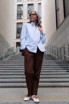business portrait of a successful senior woman with gray hair and glasses dressed in office stylish clothes on the background of the city.