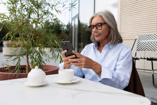 business portrait of a successful senior woman with gray hair and glasses dressed in a shirt waiting for a business lunch on the terrace.