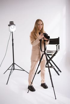 A woman producer videographer, a blonde with a camera Lumix GH5 on a tripod in the studio. Wearing a formal nude pantsuit on a white isolated background, vertical