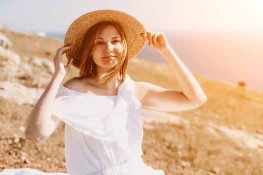 Happy woman in a white dress and hat stands on a rocky cliff above the sea, with the beautiful silhouette of hills in thick fog in the background